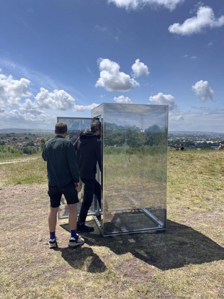 The Mirrorbox is placed on top of a hill overlooking houses and hills in the background. It is reflecting the surroundings. It is summer and the grass is dry. Two men are by the box. One is wearing a green shirt and shorts. He is holding the door open while another man in black jeans and a hooded jumper steps into the Mirrorbox. 