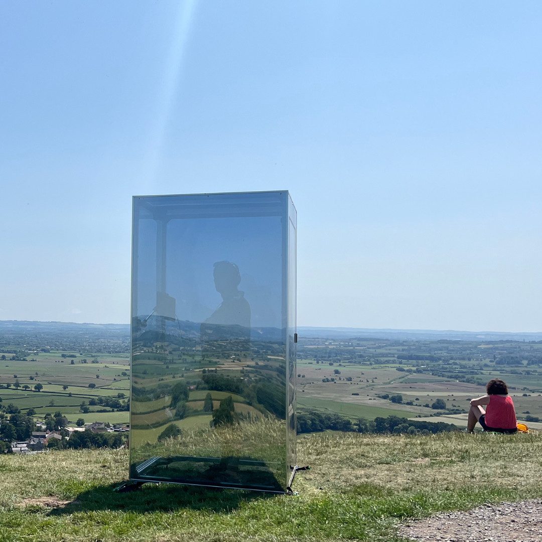 A large human-sized reflective box stands on the top of a hill. It is summer and the sky is blue. The box is reflecting the countryside around it, but you can still see the outline of a figure standing inside looking out over the fields. To one side of the box a woman in a red top is sitting on the ground.