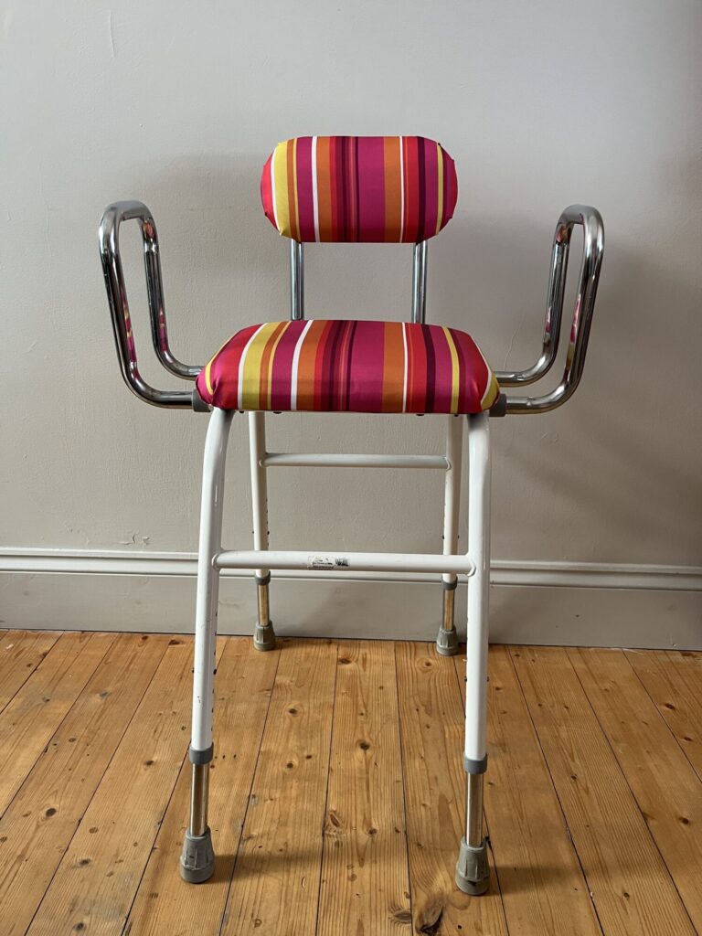 A brightly striped perching stool with four legs and a small back rest. It has two chrome arms that provide a wide seat. It is in a room with grey walls and wooden floors