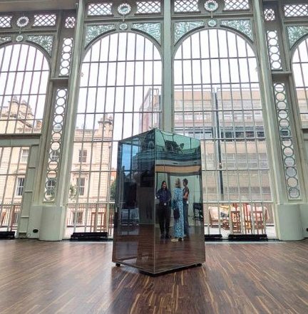 A human-sized mirrored box stands inside the Paul Hamlyn Hall at the Royal Opera House. Behind it are huge Victorian windows with Covent Garden beyond. Reflected in the Mirrorbox are people standing in a group out of the photograph. One has a blue and white dress. There is also a faint shadow of someone standing Inside the box
