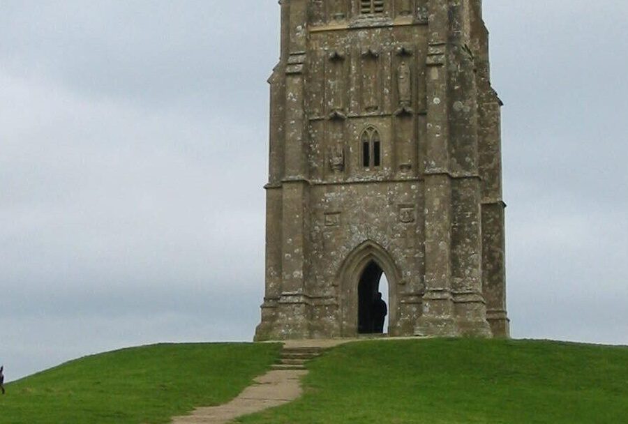 A large historic rock building at the top of a grass covered hill. There is a long path leading up to the entrance of the building which is hollowed out. There is a black dog on the left of the hill. The sky is very grey and cloudy.