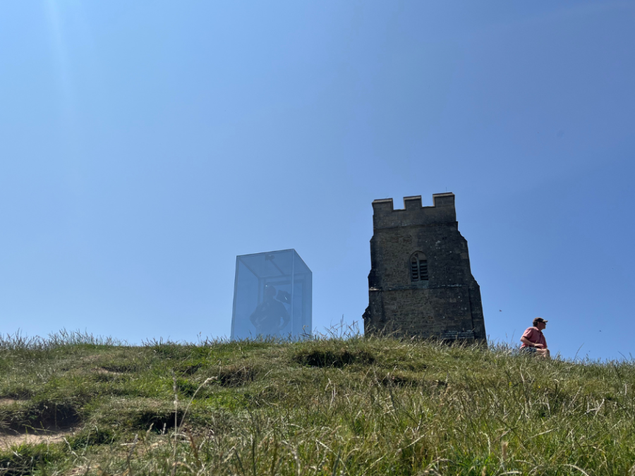It is a clear blue day. A large tower stands high above on the top of a grassy hill. Next to the tower to the left is a box. It is big enough to stand inside. In the bright sunlight you can see through it. It looks just slightly darker than the sky. The top of man in a red top can be seen walking away to the right of the box and tower