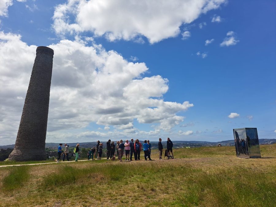 A sunny day with blue sky and clouds. A large brick chimney is on the left of the image. It is looming over a queue of people. They are waiting to enter a large mirrored box which you can see on the right of the photo. In the foreground is parched grass.